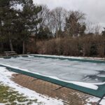Piscine enterrée couverte d'une bâche d'hivernage verte, entourée de neige dans un jardin en hiver
