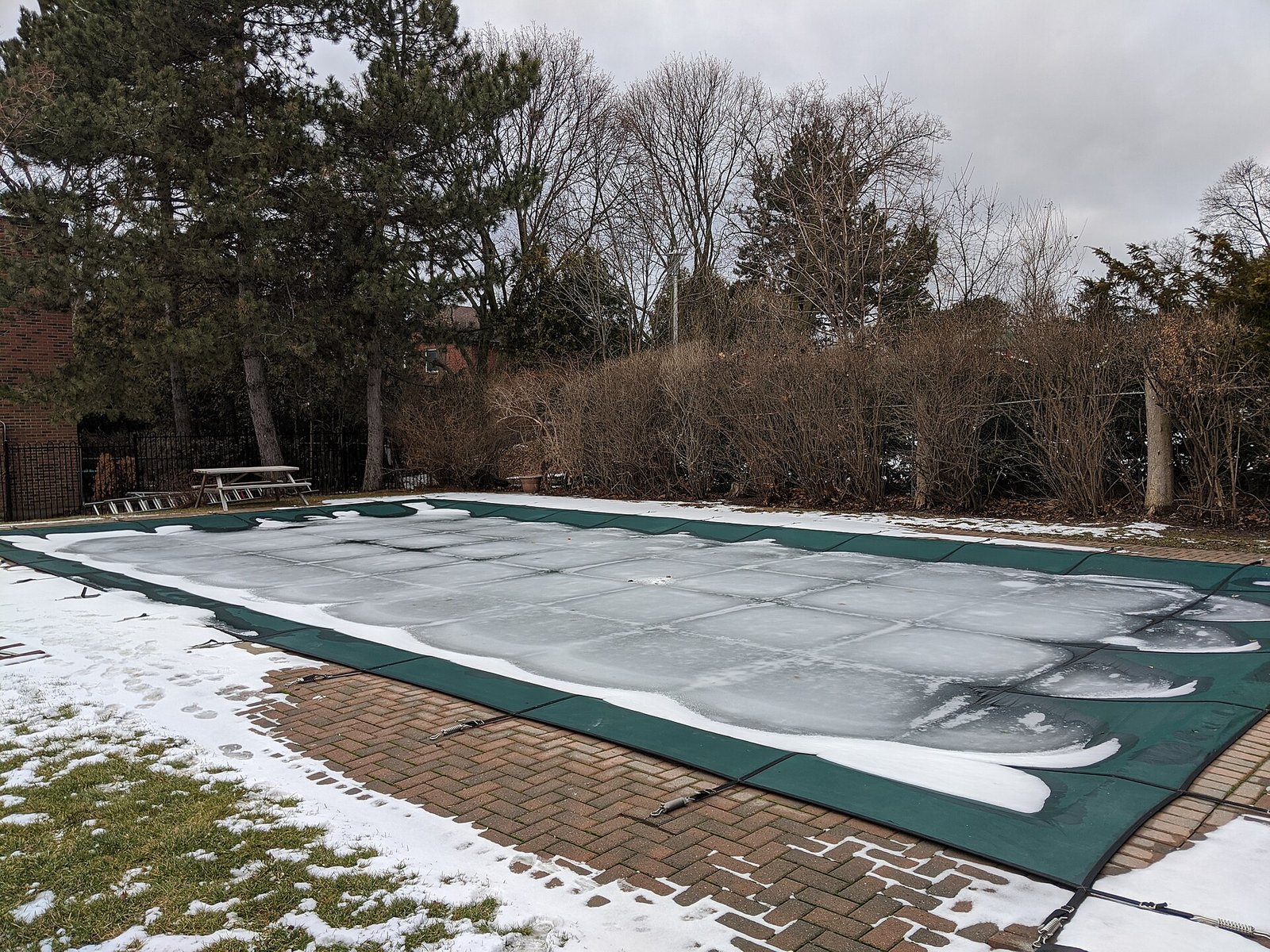 Piscine enterrée couverte d'une bâche d'hivernage verte, entourée de neige dans un jardin en hiver