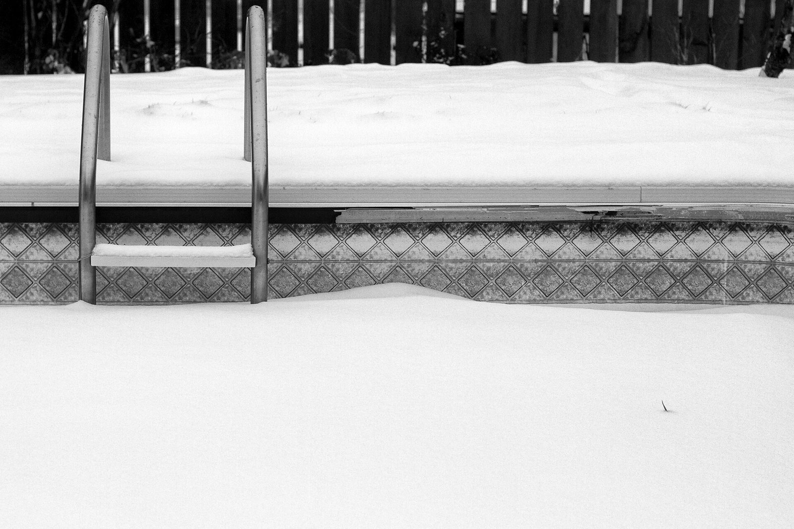 Bord de piscine et échelle sous la neige, avec la margelle et la frise de carrelage visibles